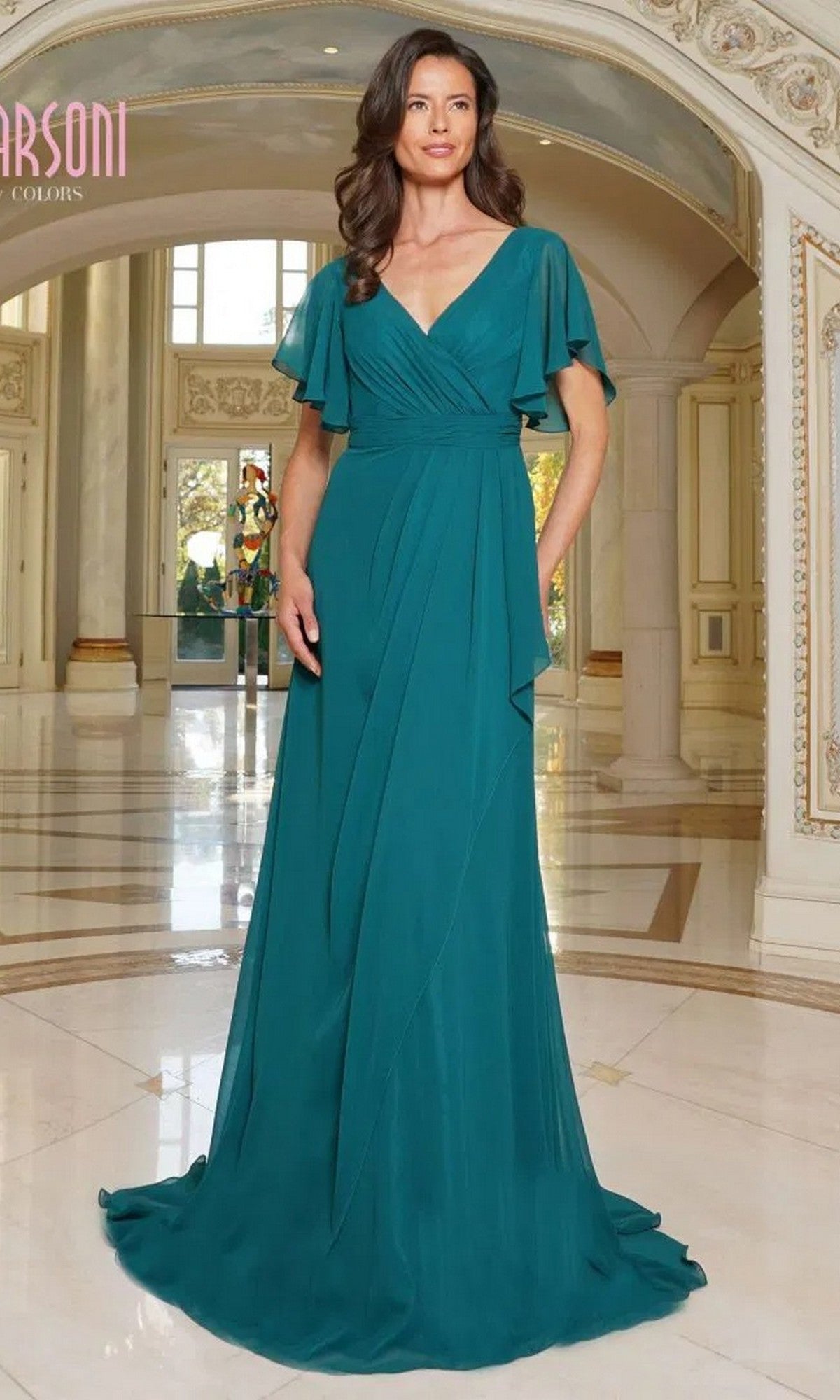 A woman in a deep green Marsoni M320 long formal dress with flutter sleeves and an a-line skirt stands in a grand hallway with marble floors and arched doorways.